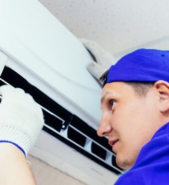 A young worker in a blue baseball cap and gloves inspects and maintains the air conditioner in the room. Portrait of a technician at work. Close-up. Mockup